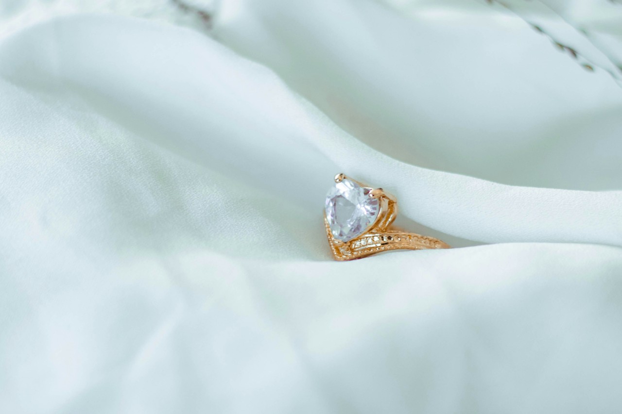 A close-up of a distinctive sculptural engagement ring with a heart-shaped center stone, delicately displayed on smooth white fabric.
