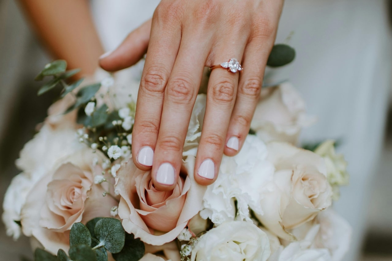 A close-up of a bride’s hand, adorned with an elegant engagement ring, atop her bouquet of roses and peonies.