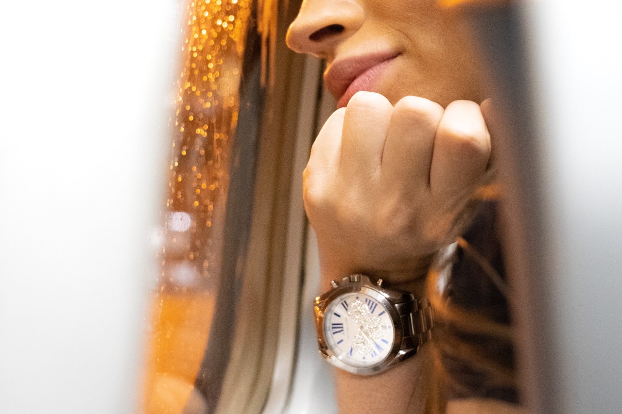 A close-up of a woman looking out of an airplane window, showcasing her metallic wristwatch with roman numerals and diamond encrusted dial.