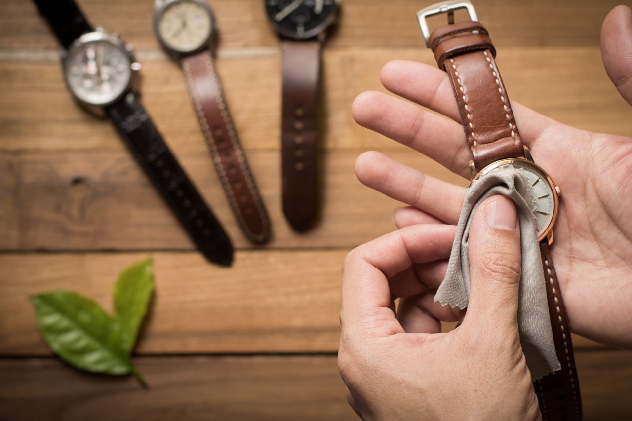 A close-up of a hands wiping brown leather wristwatch with microfiber cloth with 3 others laying on a wooden surface in the background