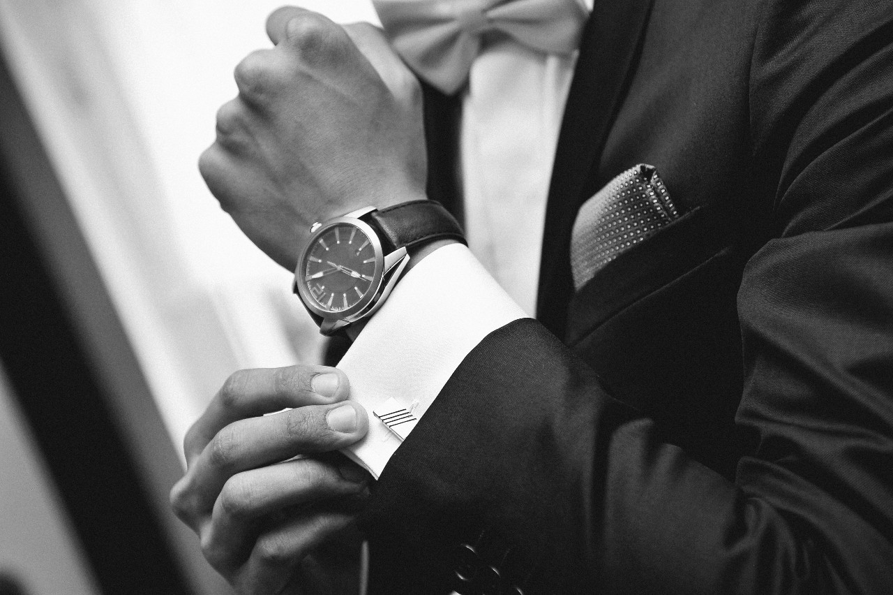 A black and white picture of a man in a suit fixing his cufflinks, showcasing a leather wristwatch with black dial.