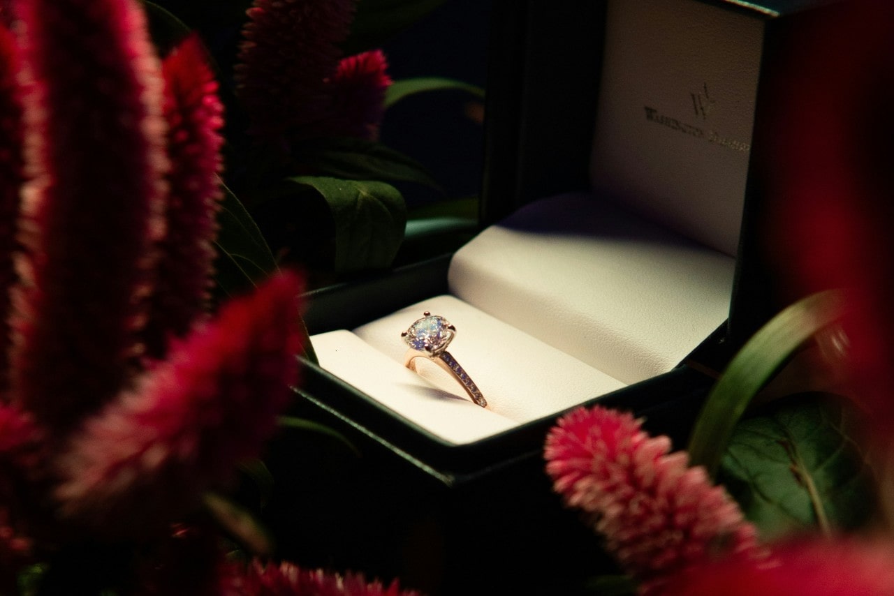 A close-up of a diamond side stone engagement ring in a black ring box surrounded by flowers.