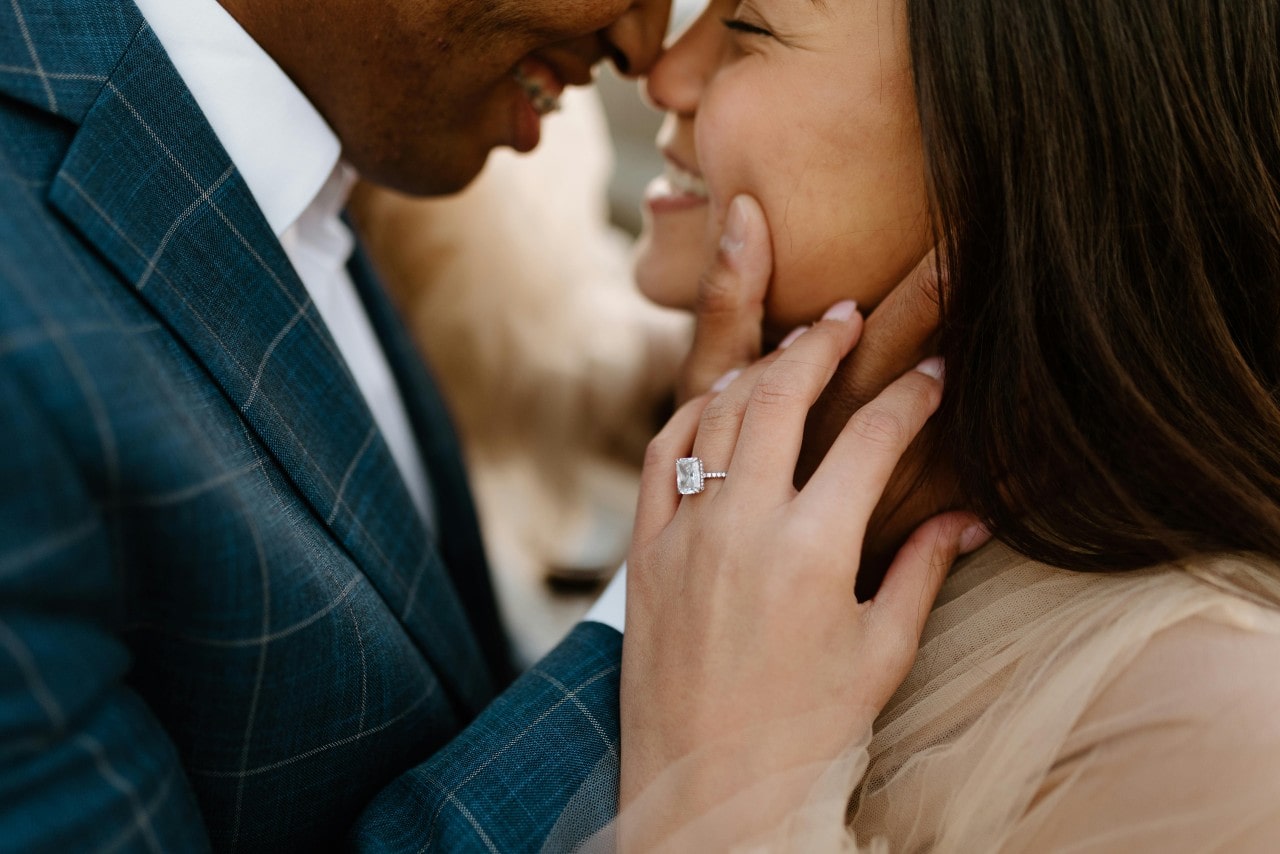 A close-up of a man and woman embracing— the woman wearing a white gold engagement ring with an emerald cut diamond.
