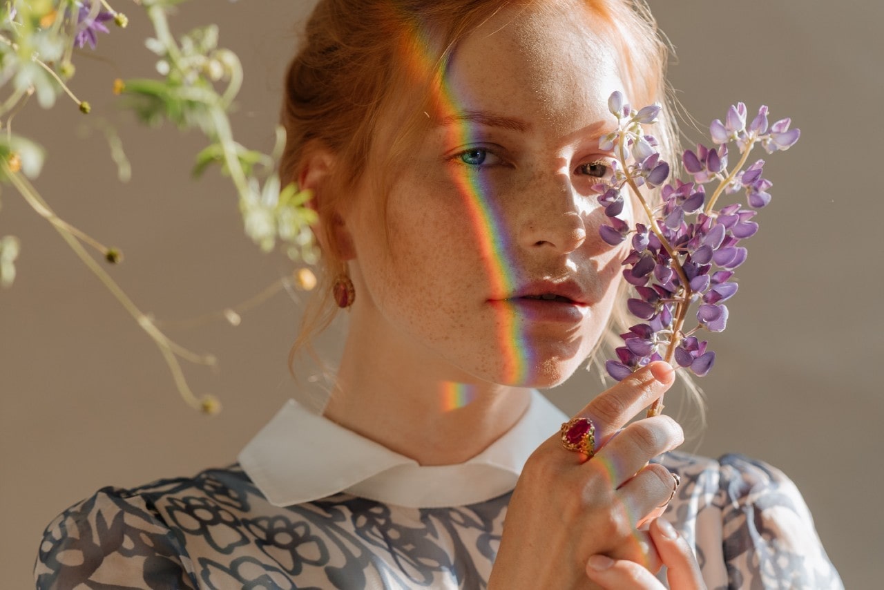 A woman holding a flower wearing gemstone jewelry with a rainbow across her face.