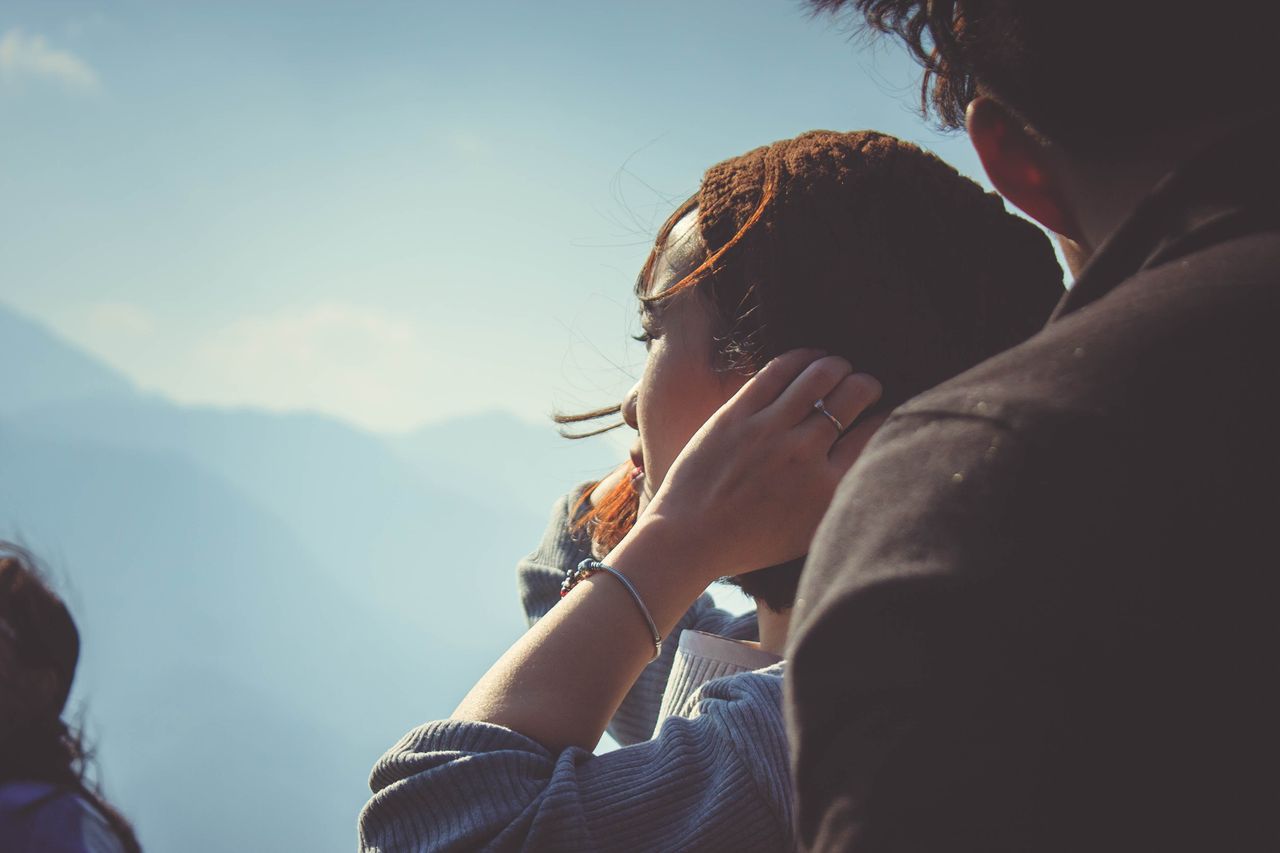 A woman with long hair, facing away and adjusting it beside another person, flaunting a sparkling ring and bracelet against misty mountain vistas.