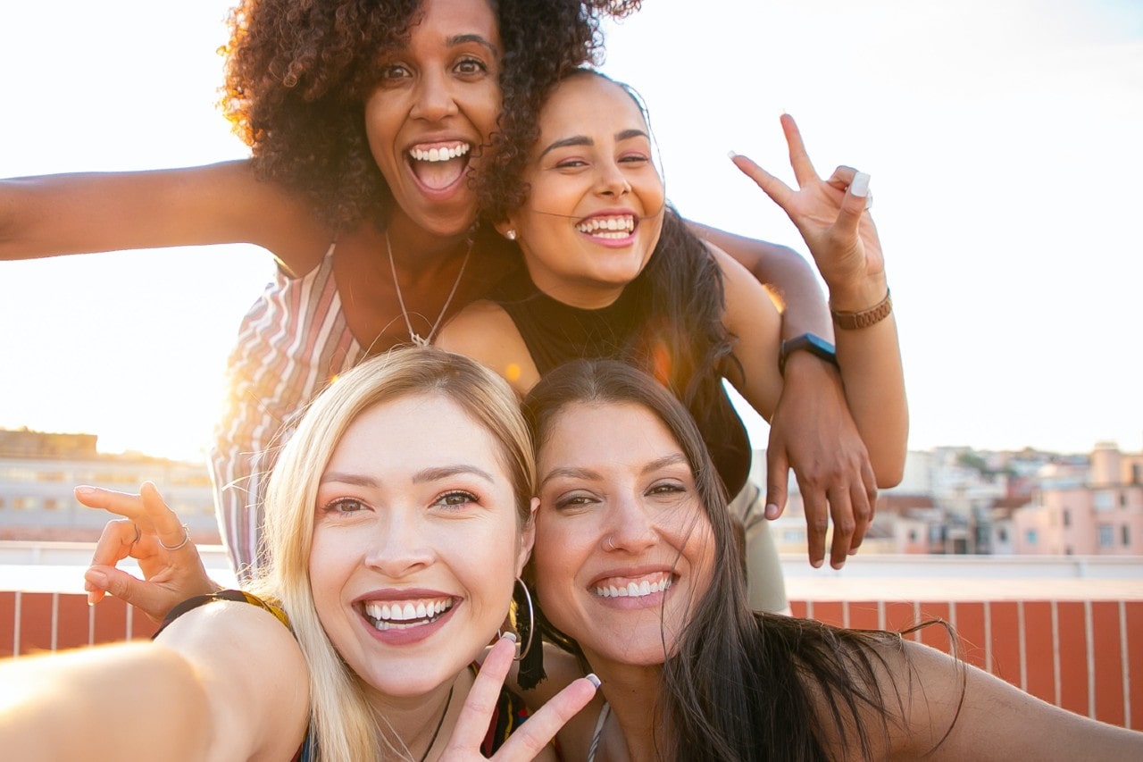Four friends wearing fashion jewelry, joyfully posing for a selfie on a sunny rooftop.