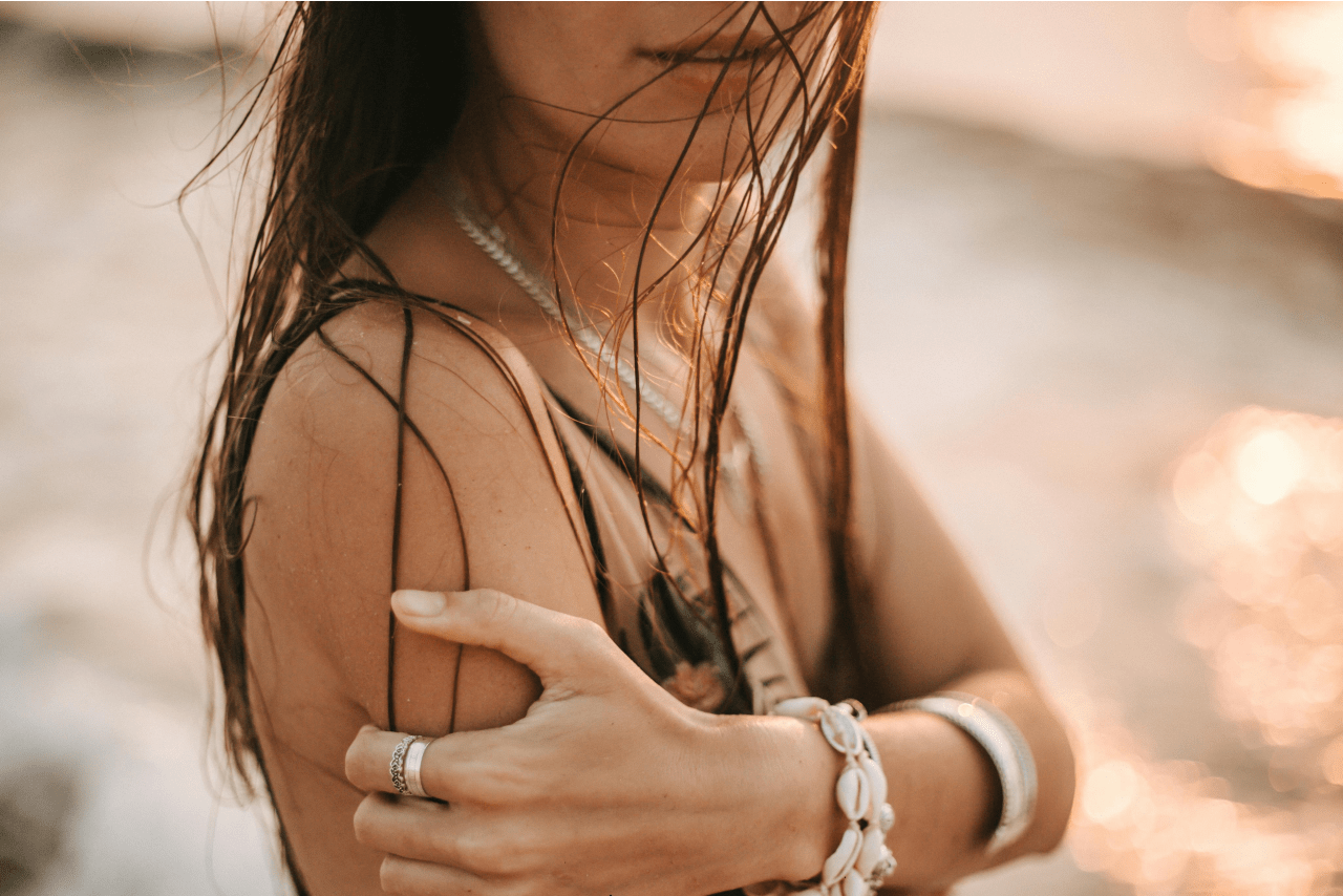 A woman with wet hair holds her arm by the beach at sunset wearing shell jewelry.