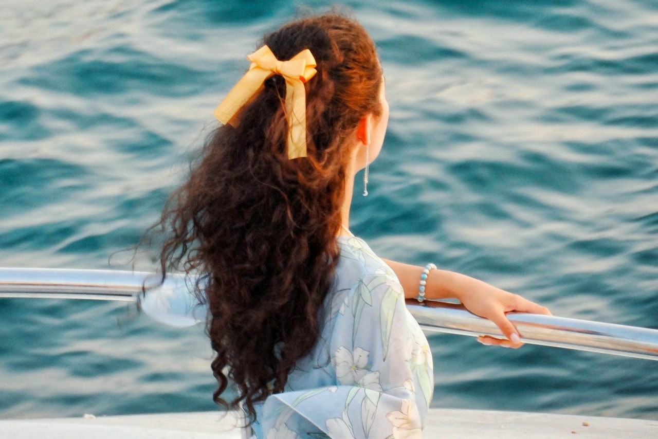 A woman with long, curly hair tied with a yellow bow gazes at the ocean from a boat.