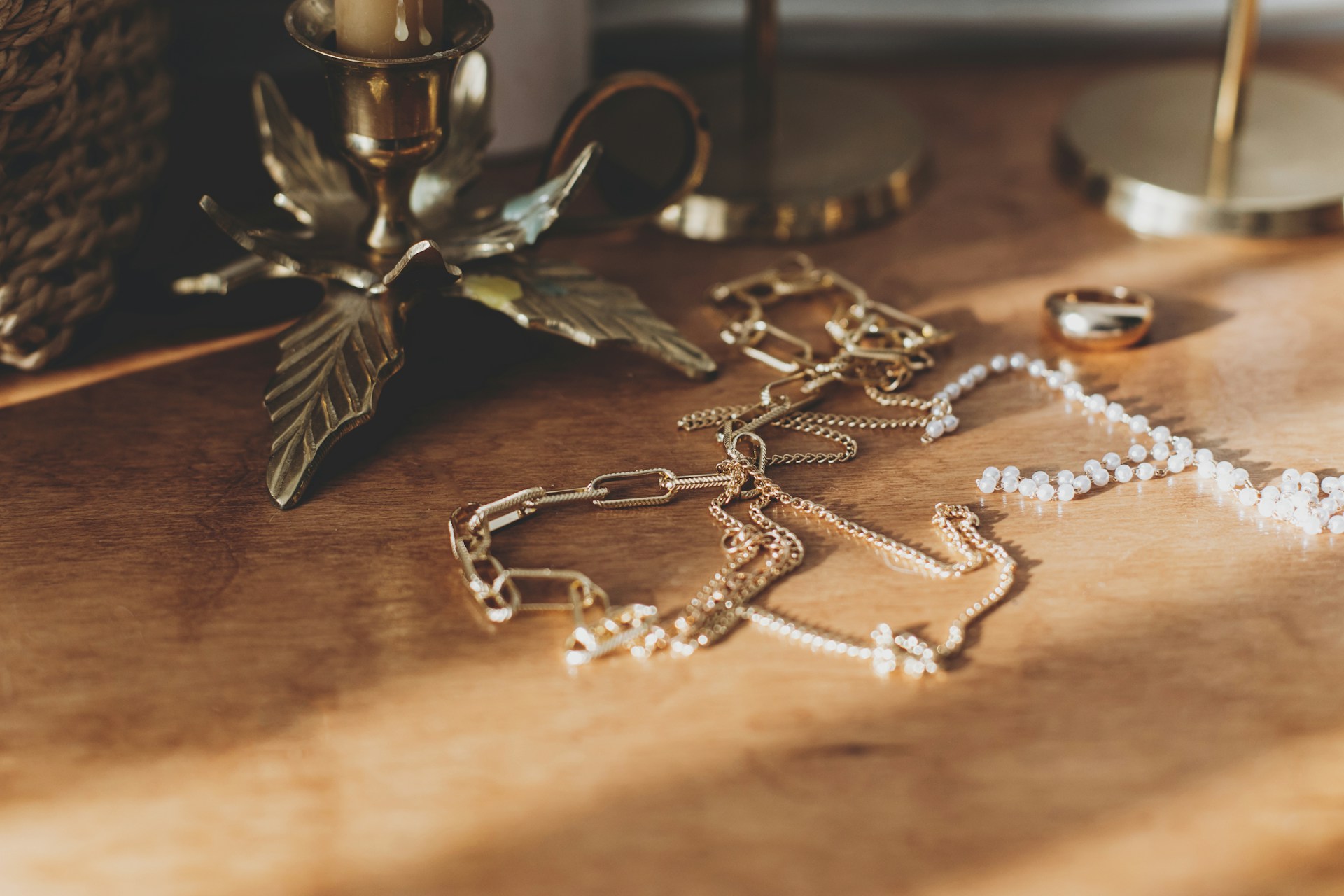 Gold and pearl necklaces are aesthetically arranged on a wooden surface beside a brass candle holder with a leaf design.