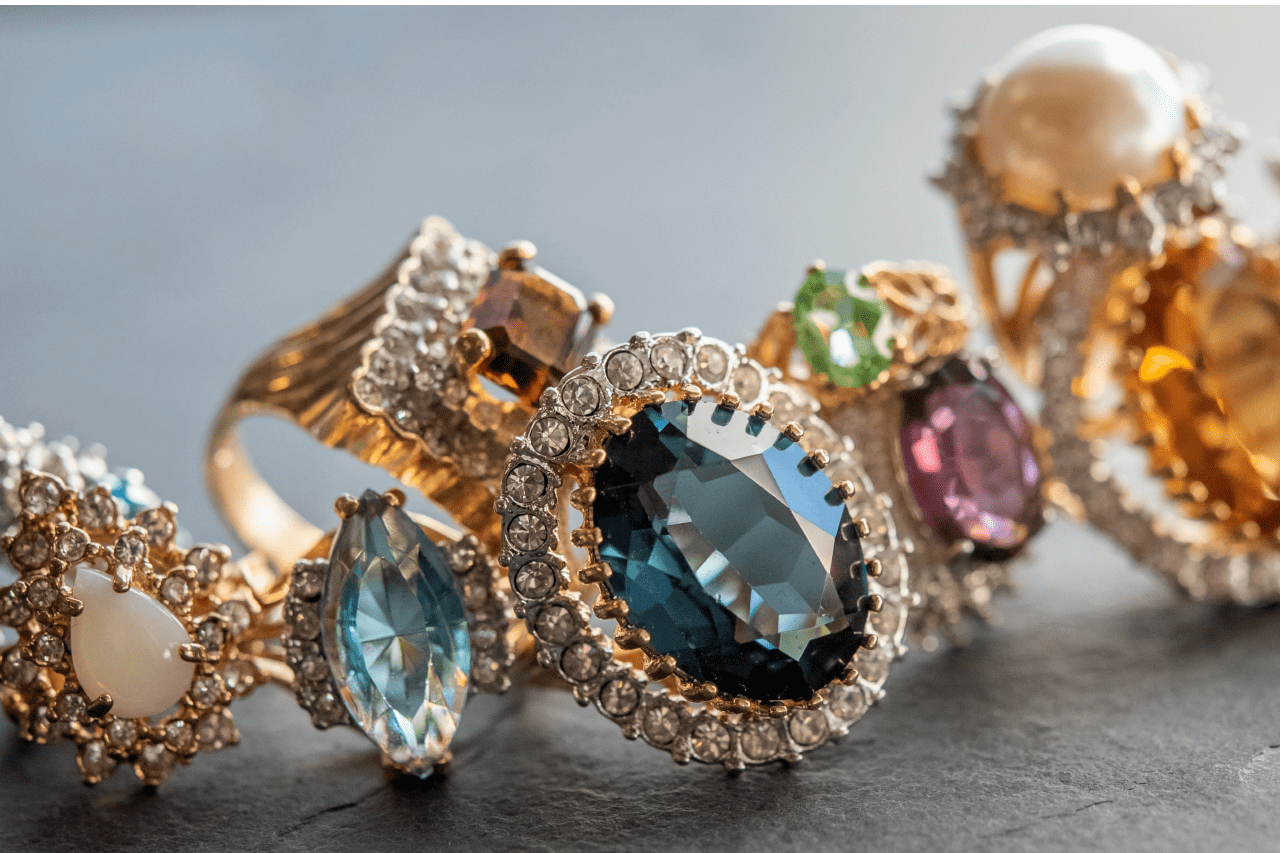 A close-up of vintage-style gemstone rings in gold settings, featuring a variety of stones like sapphire, opal, citrine, and amethyst, all accented with diamond halos.