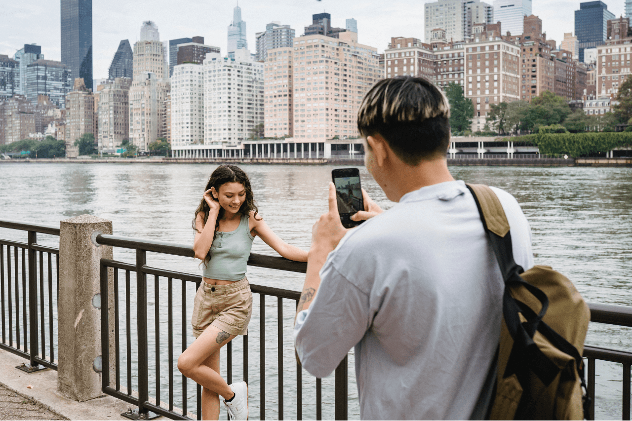 A woman poses by a lake with a big city behind her while a man takes a cell phone picture.