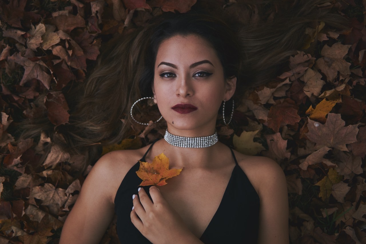 A woman wearing a black dress with silver hoops and a diamond choker lies in a pile of leaves.