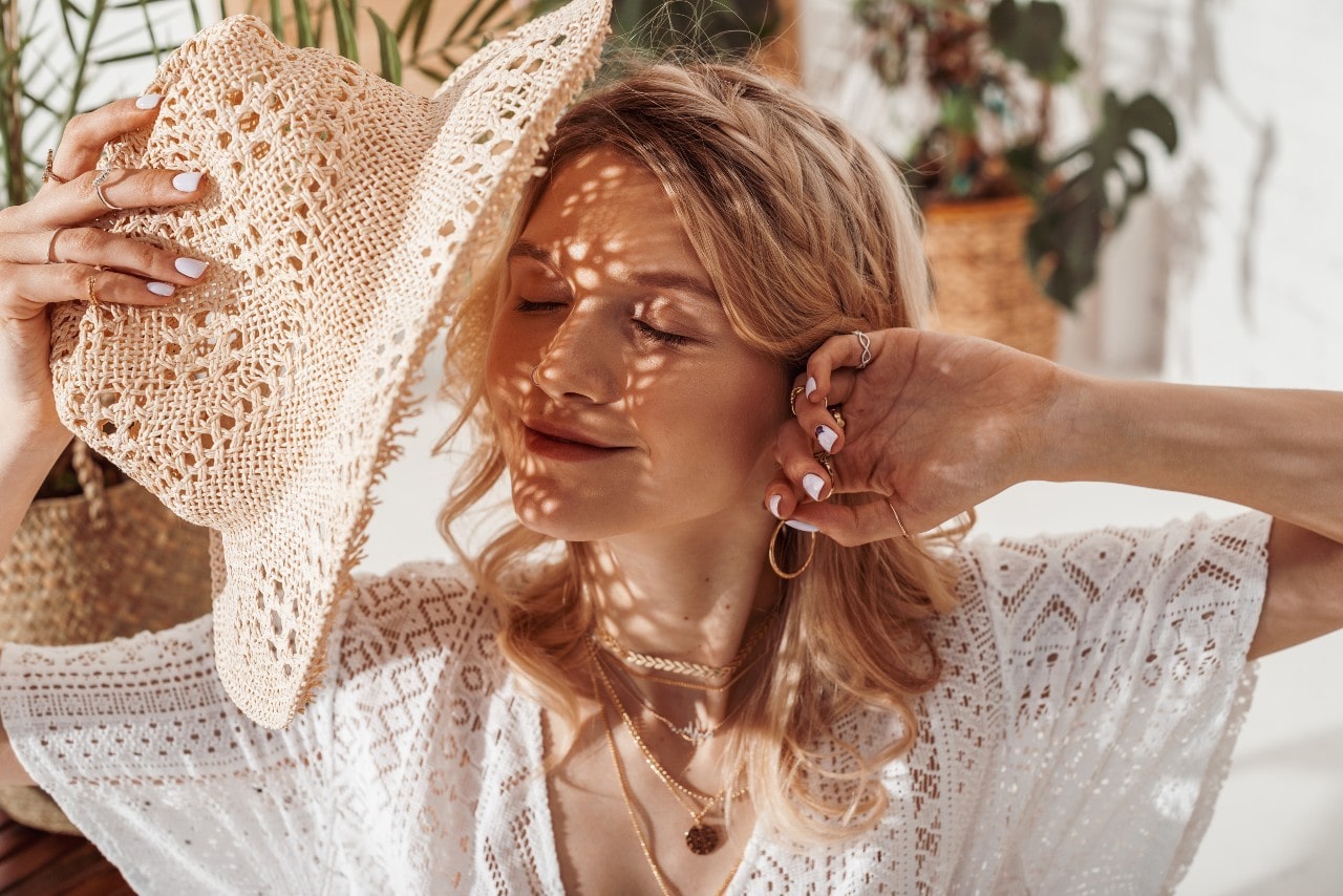 Woman shading her face with a straw hat. Sh has multiple gold necklaces, rings, and gold hoop earrings.