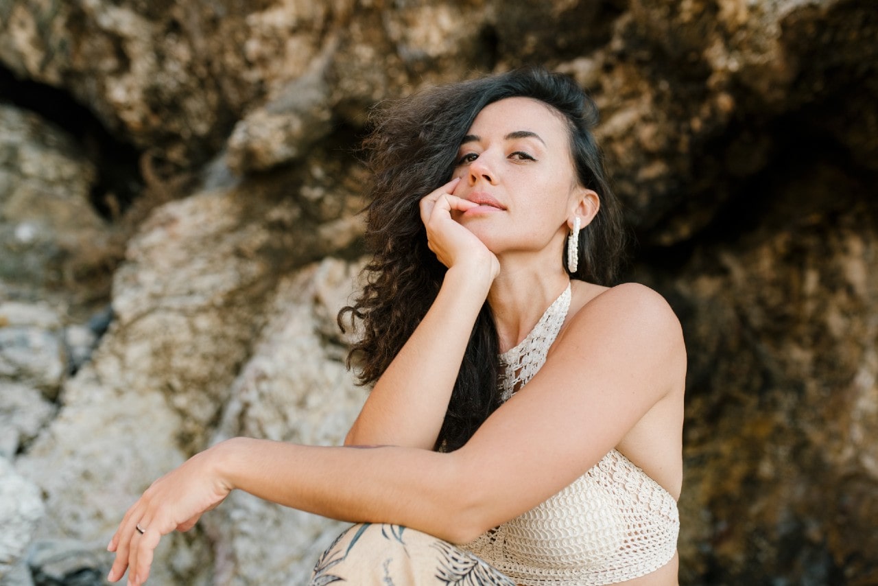A woman with curly dark hair in a crochet halter top wears diamond earrings and poses outdoors against a rocky beach background.