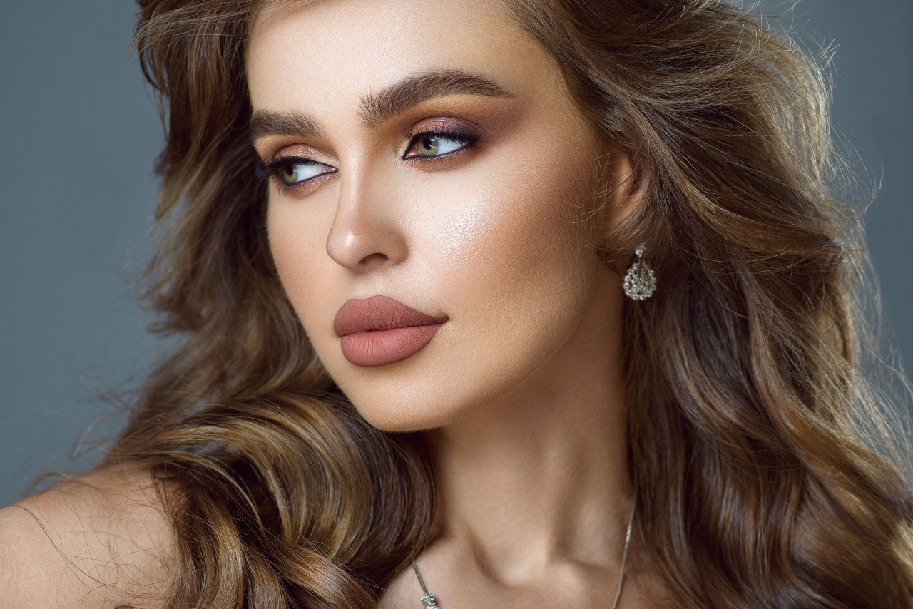 A close-up of a woman with long wavy brown hair, wearing diamond earrings and a delicate pendant.