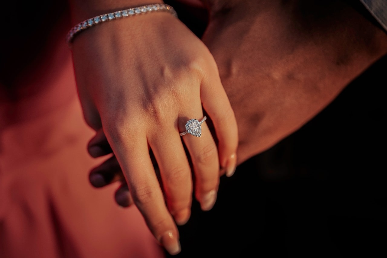 A white gold halo pear shaped engagement ring and diamond tennis bracelet on a woman’s hand that is on top of the man’s hand.