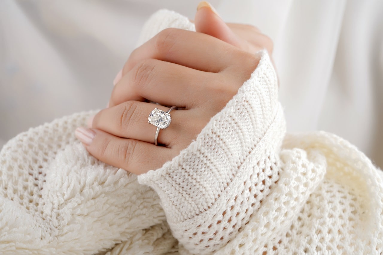 A close-up of a woman&rsquo;s hand, wearing a white knit sweater and showcasing her solitaire engagement ring.