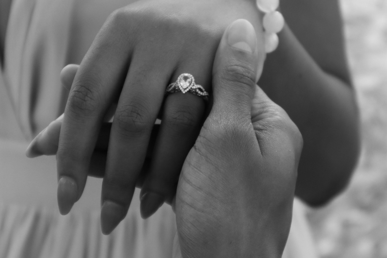 A close-up black and white image of a man holding a woman&rsquo;s hand softly, showcasing her pear-shaped engagement ring.