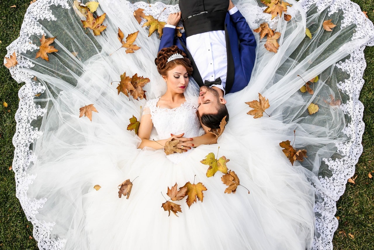 A bride in a white gown and groom in a blue suit lie on grass surrounded by autumn leaves, gazing lovingly at each other.