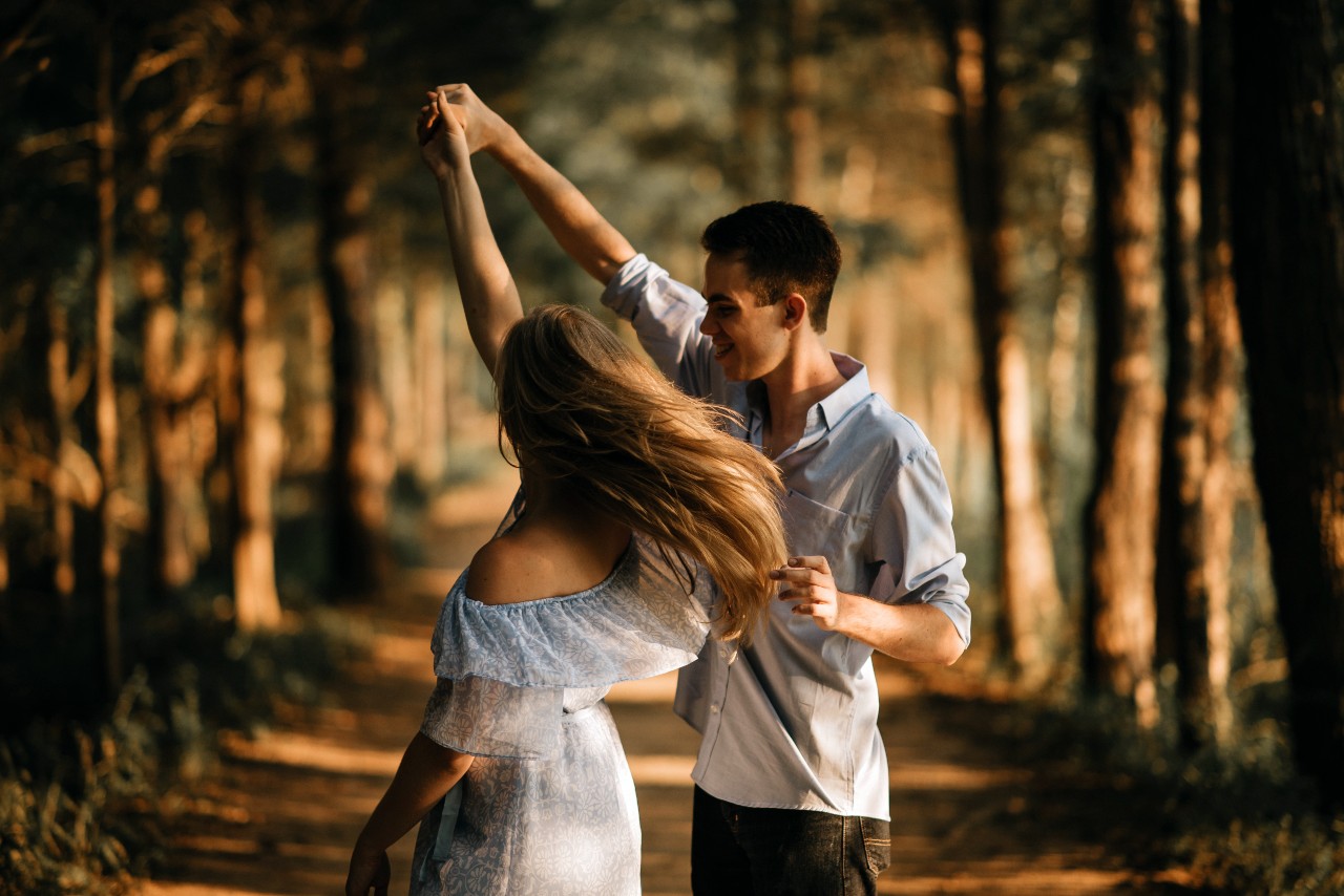 A couple dances joyfully on a sunlit forest path, the woman’s light off-shoulder dress flowing as the man twirls her.