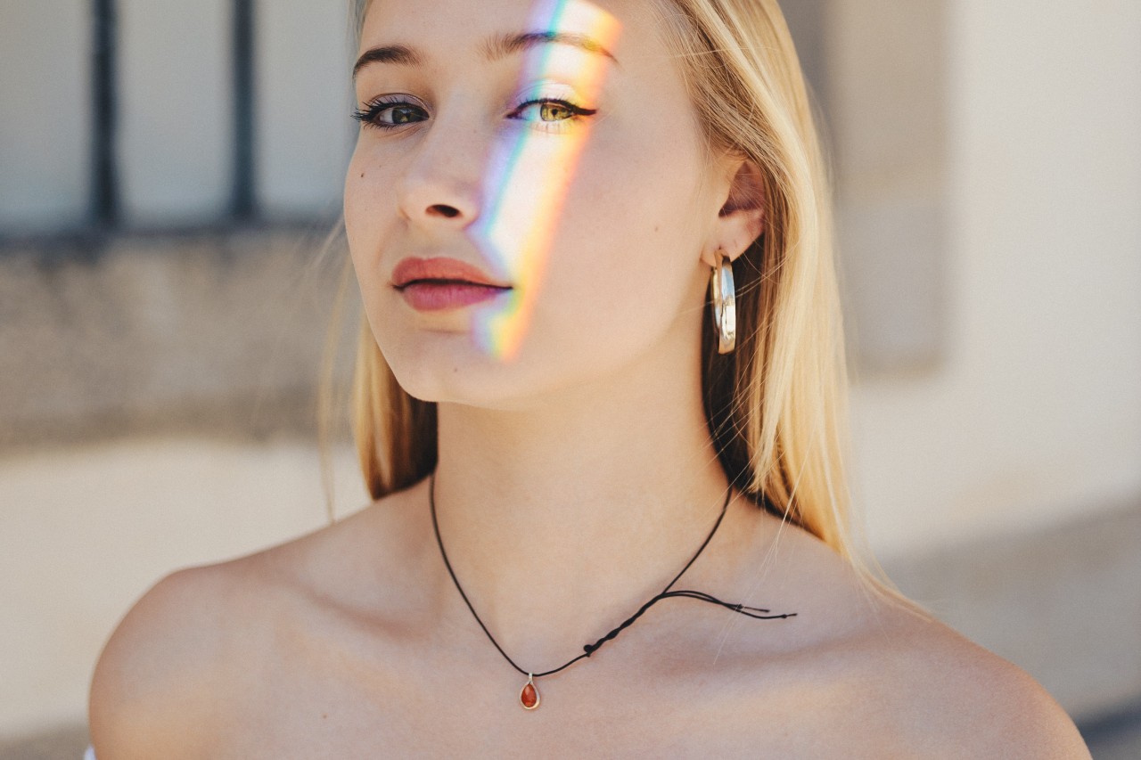 A young woman posing under a prism rainbow wears a gemstone necklace.