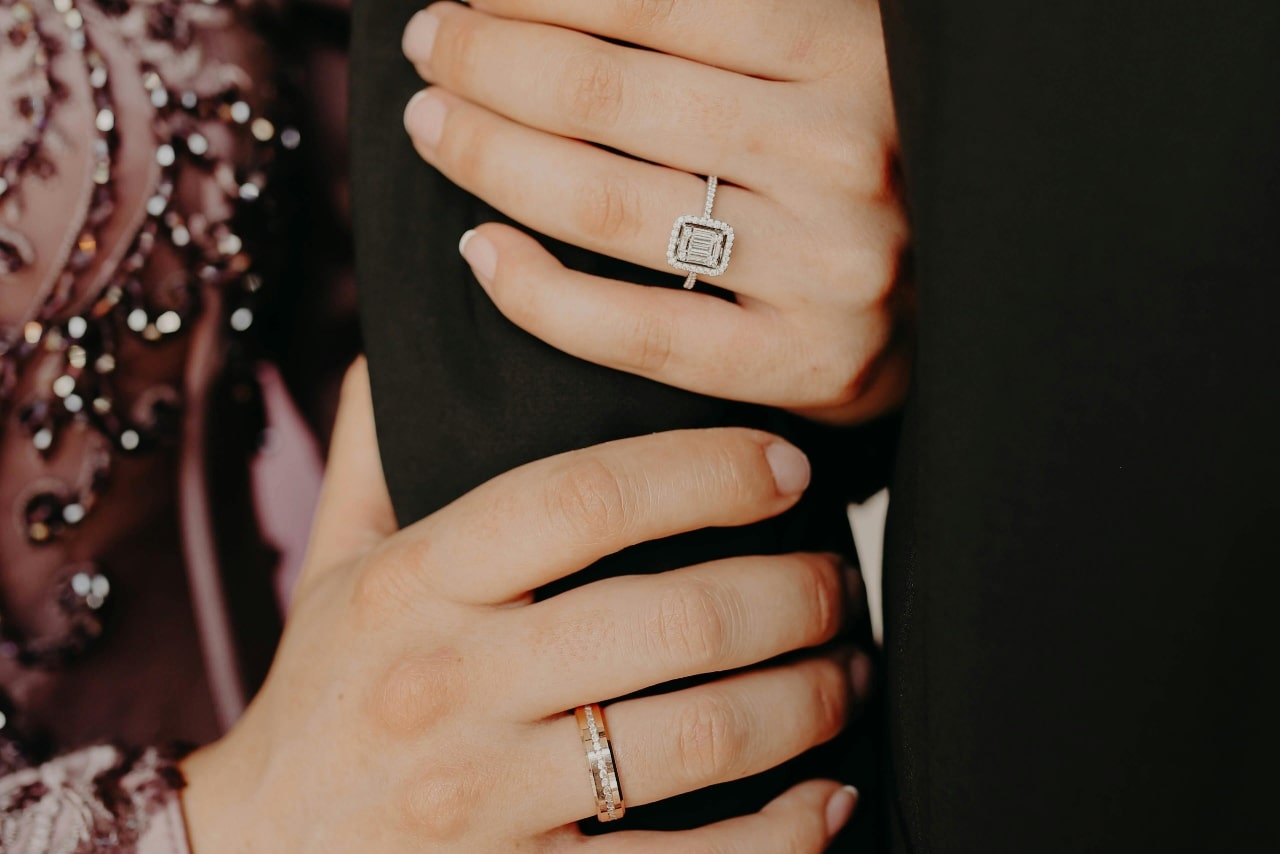 A close-up of a person&rsquo;s hands, one adorned with an emerald cut halo ring and the other a subtle wedding band, holding their partner&rsquo;s arm.