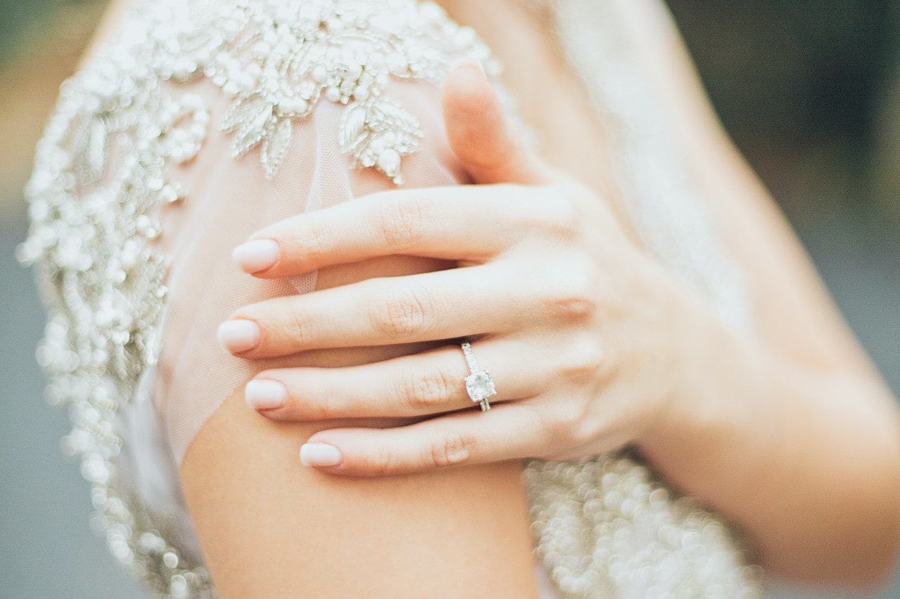 Close-up of a bride’s hand wearing a side stone diamond engagement ring, resting on her shoulder over a lcaed gown.
