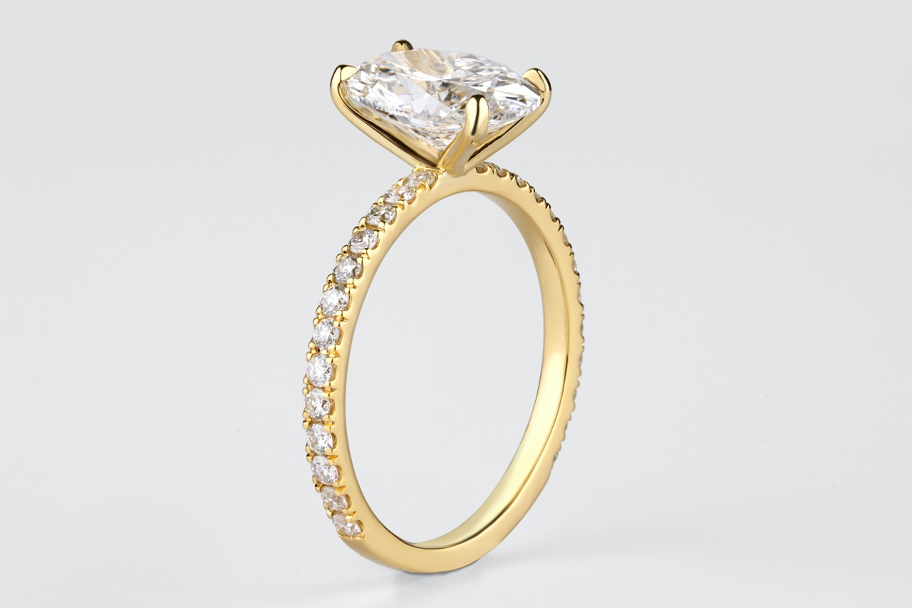 Gold engagement ring featuring a large cushion cut diamond in a four-prong setting, with a pave band adorned with smaller diamonds.