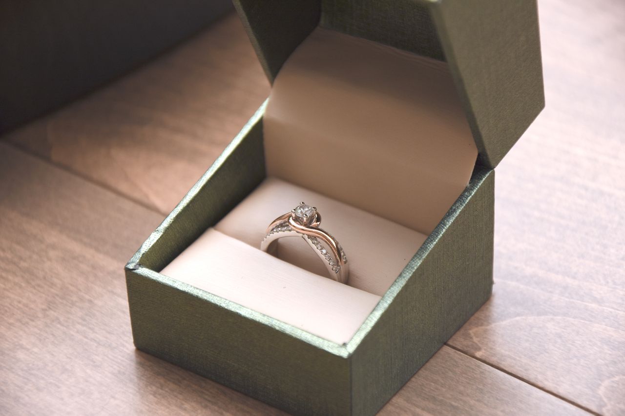 A mixed-metal engagement ring inside a green ring box resting atop a wooden table.