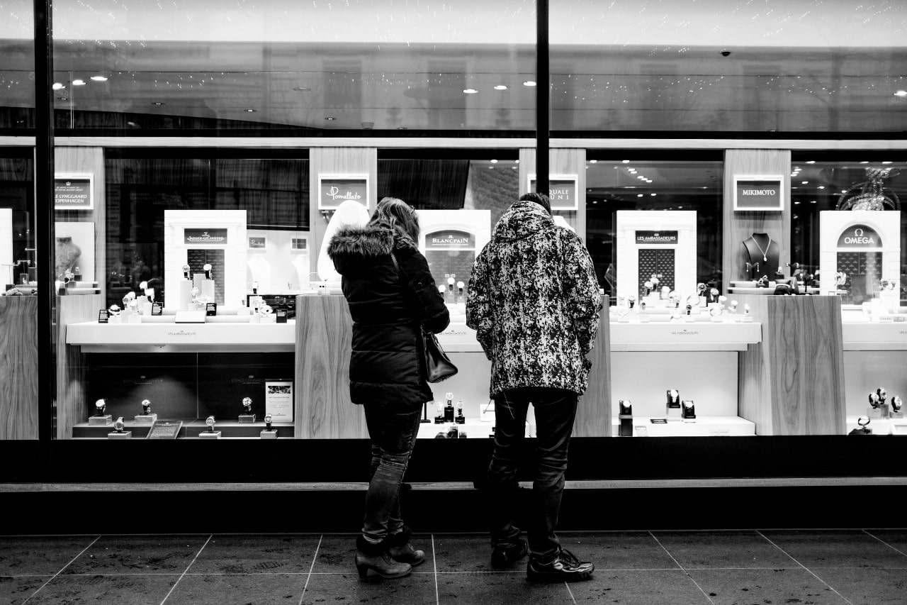 A black-and-white image captures two warmly dressed shoppers admiring jewelry through a store window.