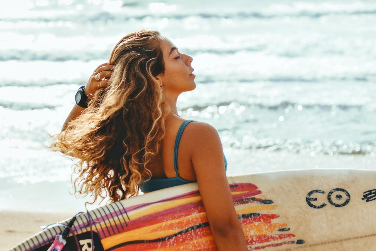 A woman about to go surfing and is wearing a watch along with a couple pieces of fashion jewelry