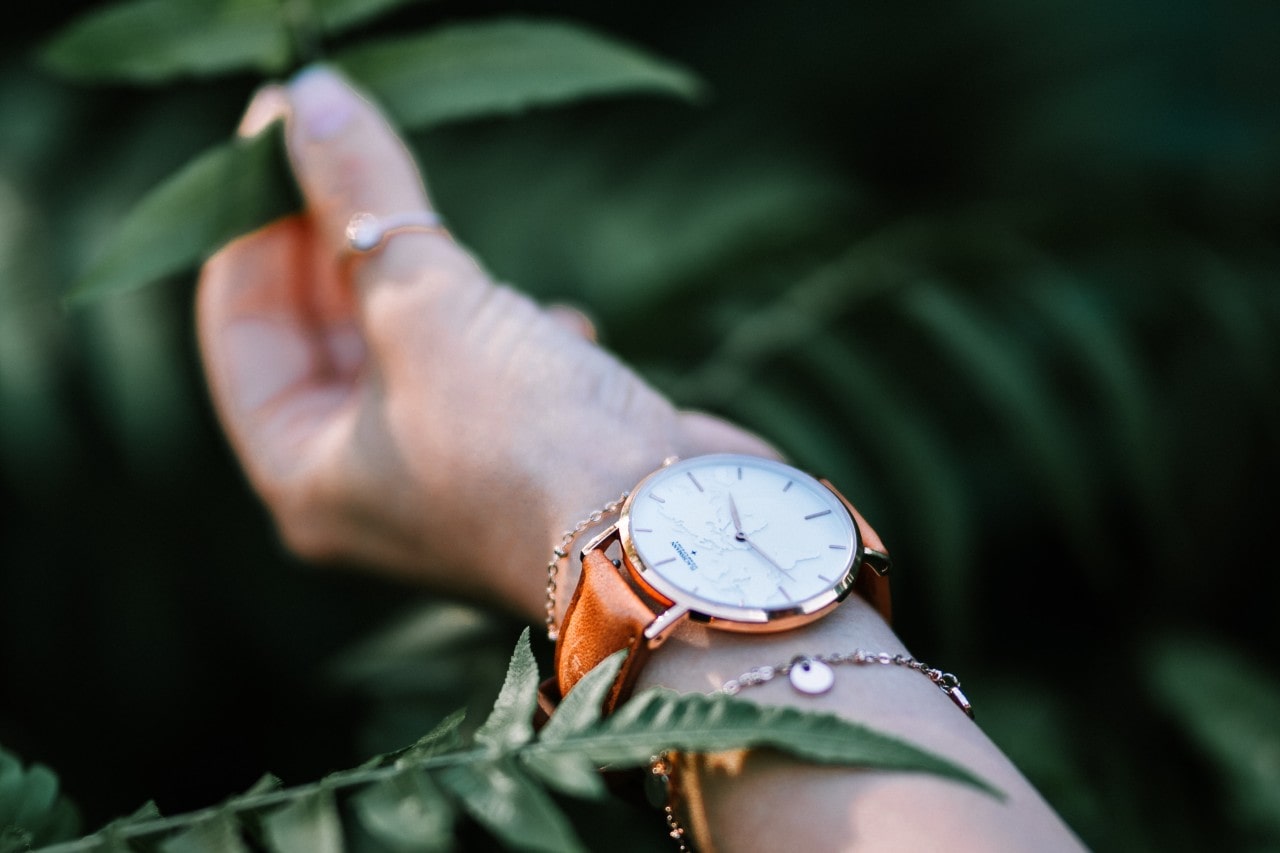 Tan leather band with gold case watch in between two gold chain bracelets on a woman’s wrist reaching out toward leaves