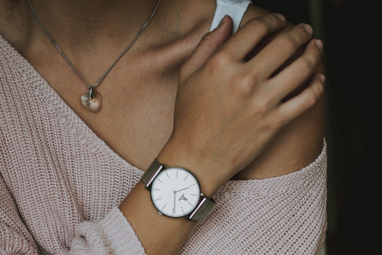 Woman holding her shoulder while wearing a slouchy pink sweater, a watch with a mesh band, and a quartz heart pendant necklace