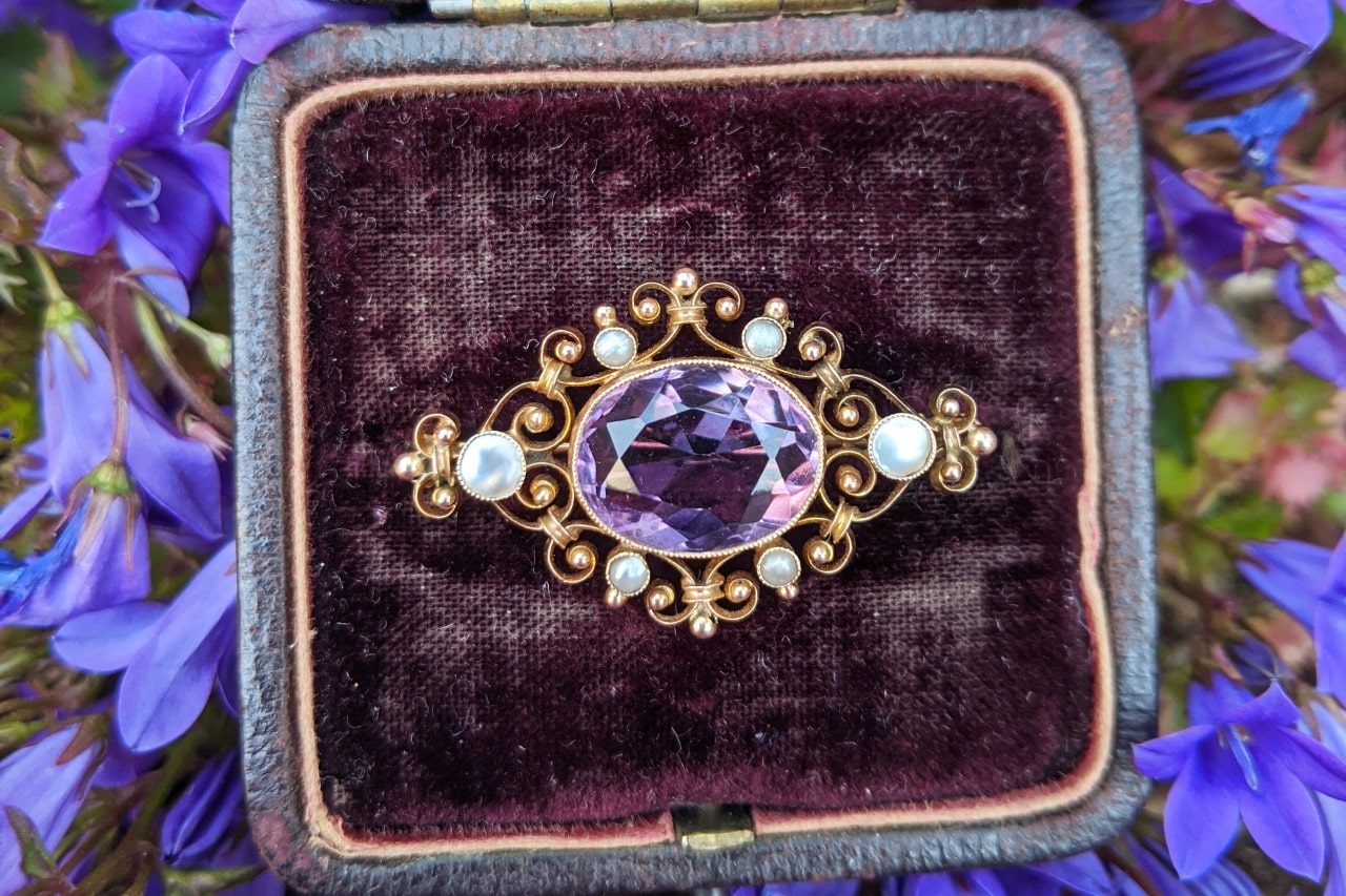 A vintage gold brooch with a purple center stone and small white pearls, placed on a dark velvet cushion surrounded by purple flowers.