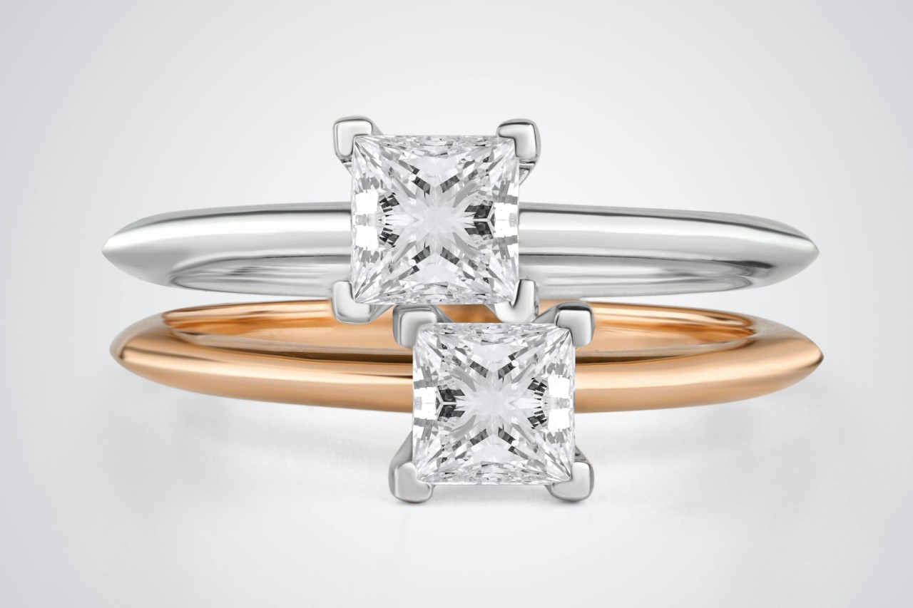 A close up of two solitaire princess cut diamond engagement rings, one with white gold band and another with yellow gold against a white background.