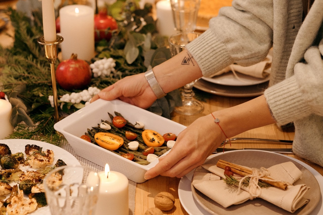 A woman places a dish on the Thanksgiving table wearing bracelets and a cardigan.