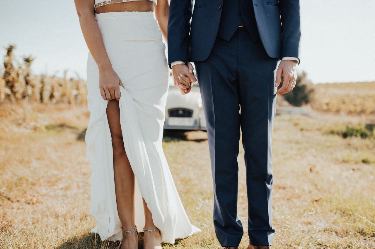 a bride and groom standing outside in front of a car, the bride holding the hem of her dress.