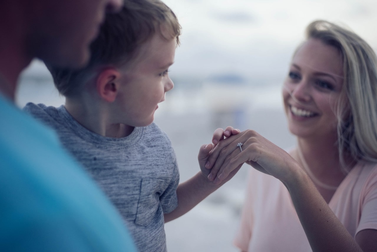 A little boy holding the hand of his mom who has a new halo engagement ring after the heartfelt proposal by the man holding the little boy at sunset