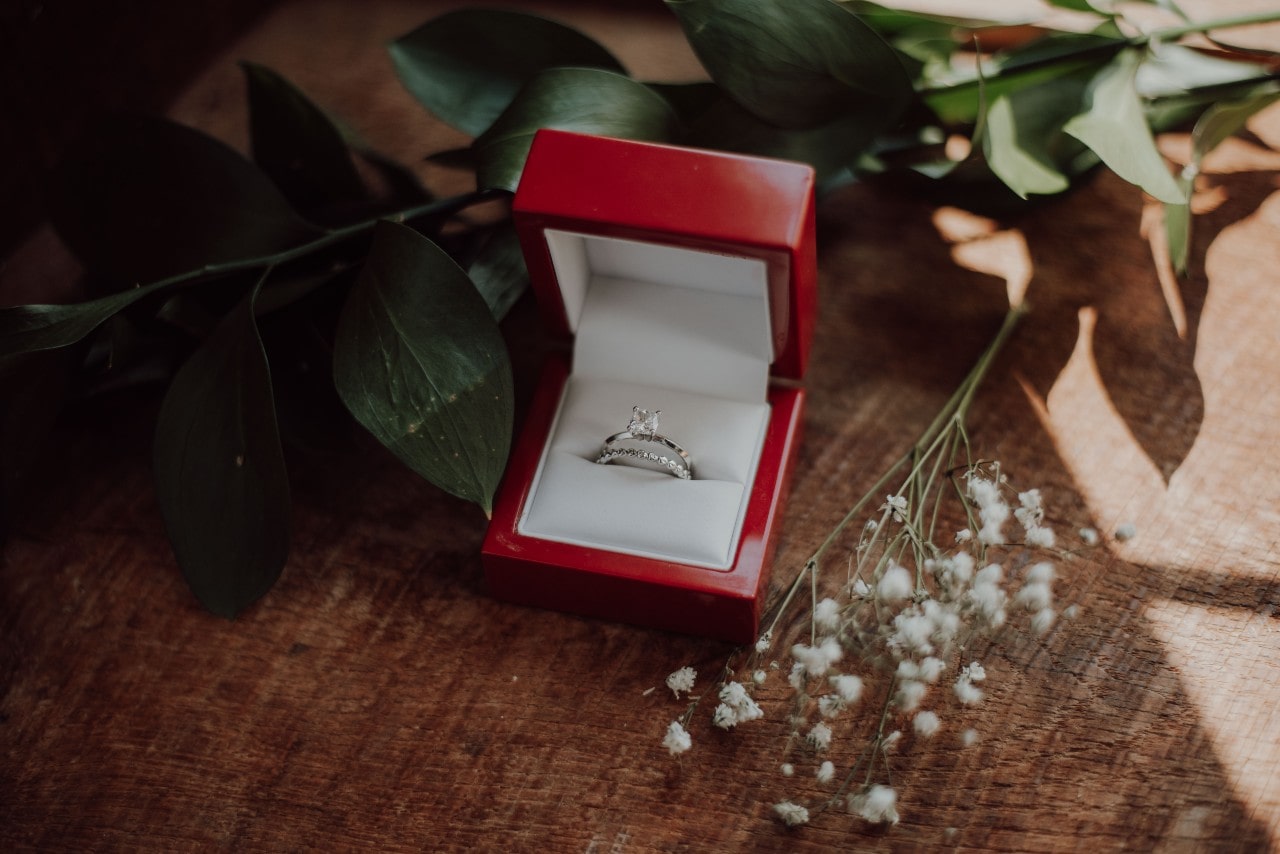 an engagement ring and wedding ring in a red box on a table next to flowers