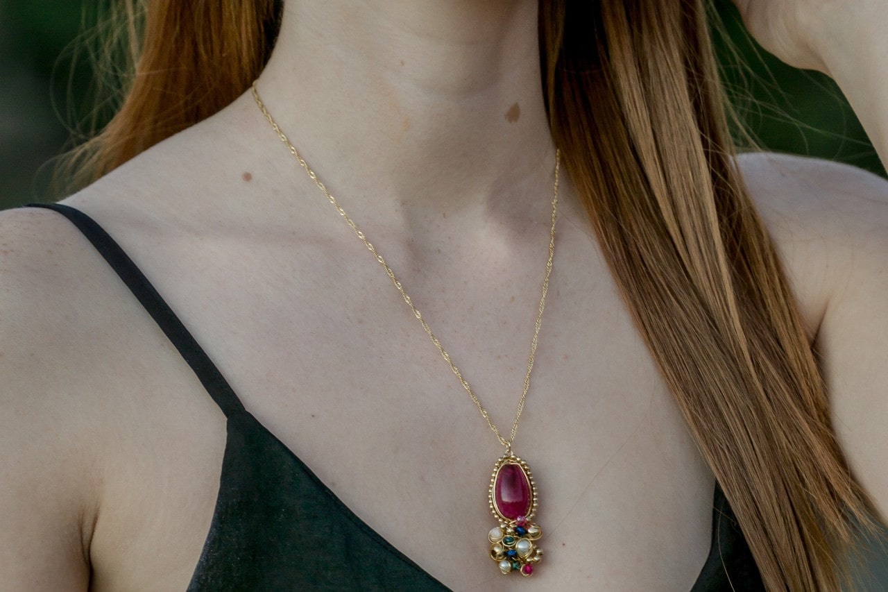 A close-up of a woman wearing a delicate yellow gold ruby gemstone pendant accented by diamonds, pearls, and colorful gemstones.