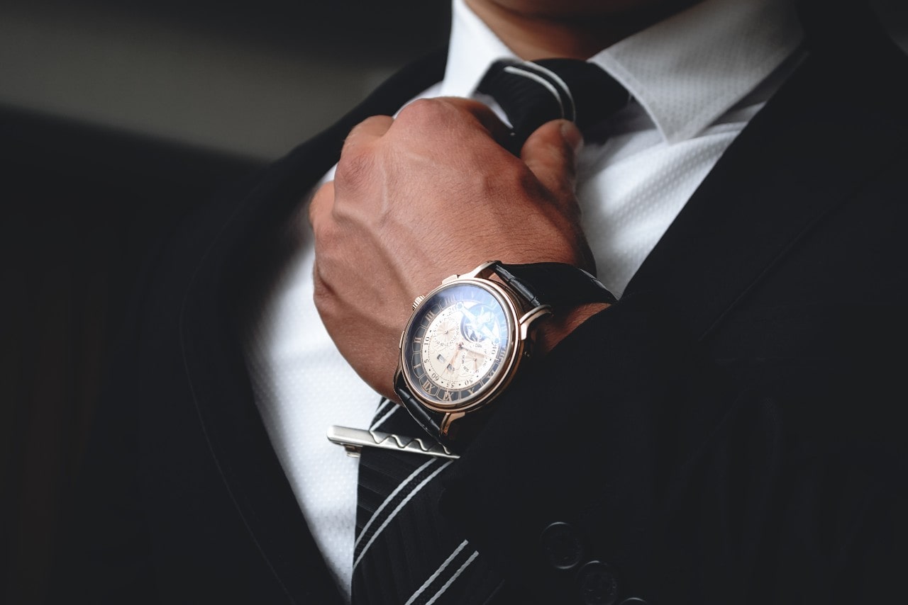 a man’s hand adjusting his black and white striped tie and wearing and intricate watch