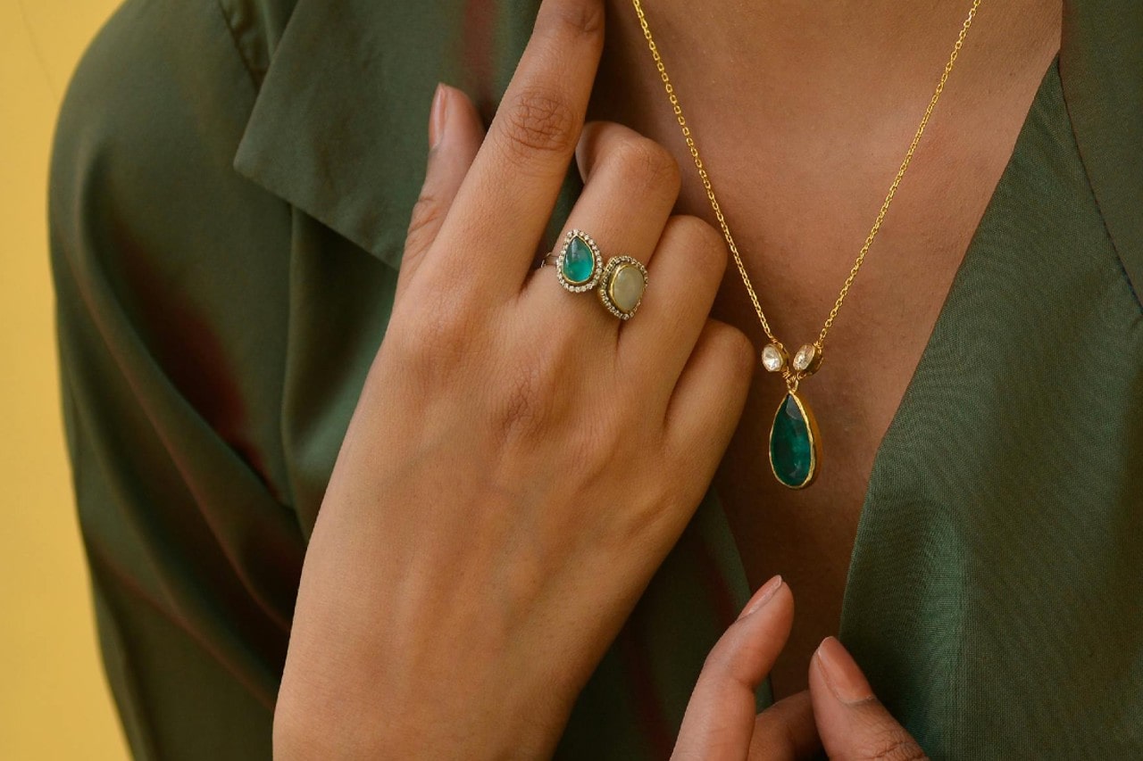 A close-up of a person in a green shirt wearing a teardrop emerald pendant and a matching green gemstone cocktail ring.