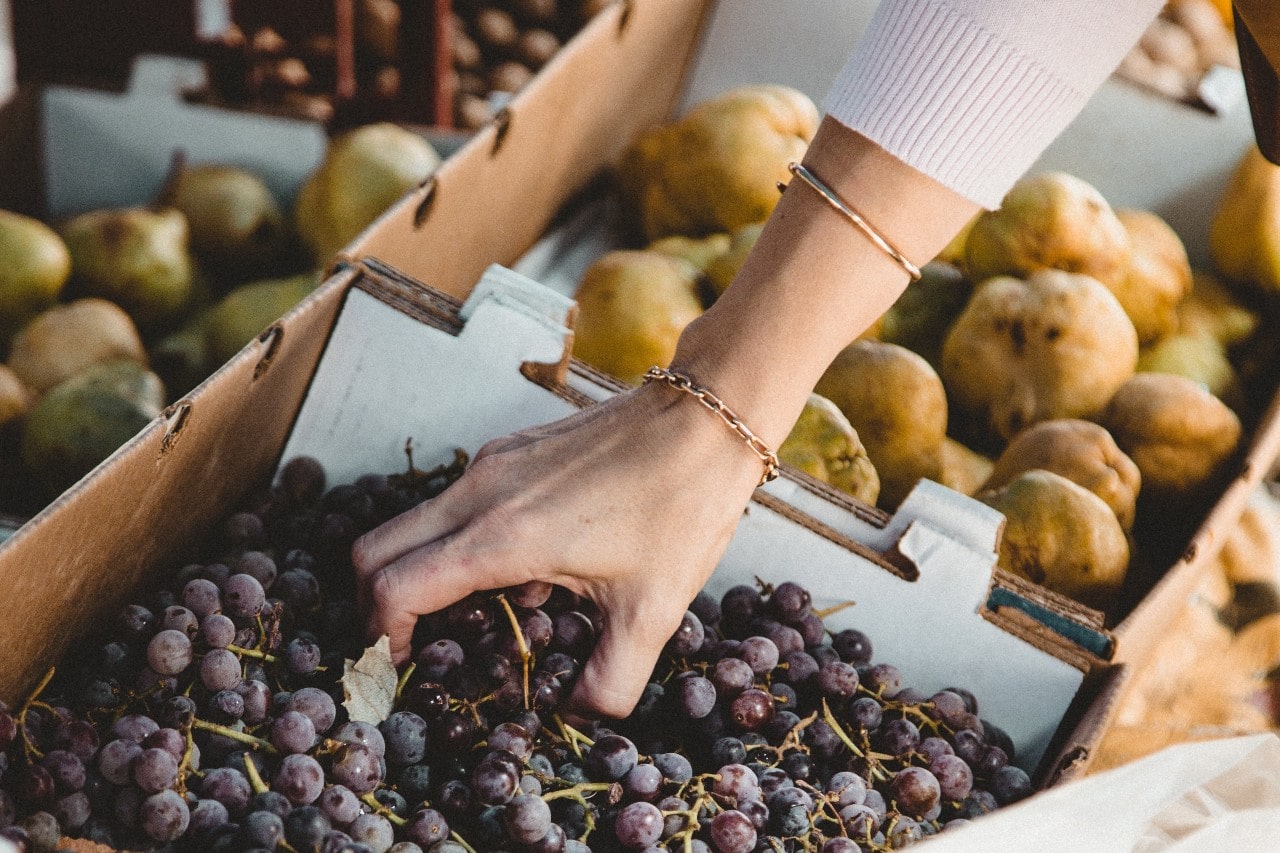 A woman wearing gold bracelets reaches for a bundle of black grapes at a grocery store.