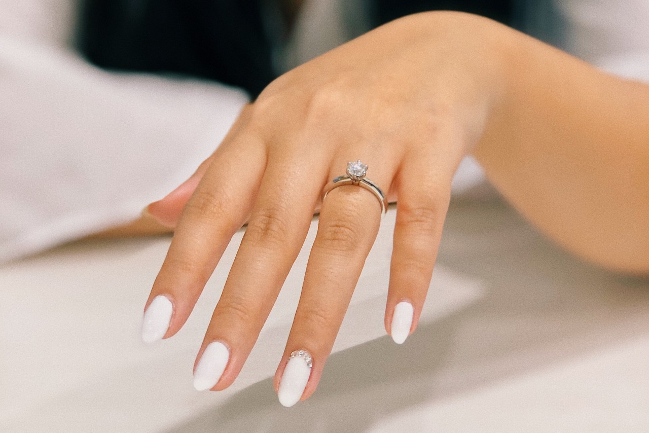 a woman’s outstretched hand, with white nails, wearing a solitaire engagement ring