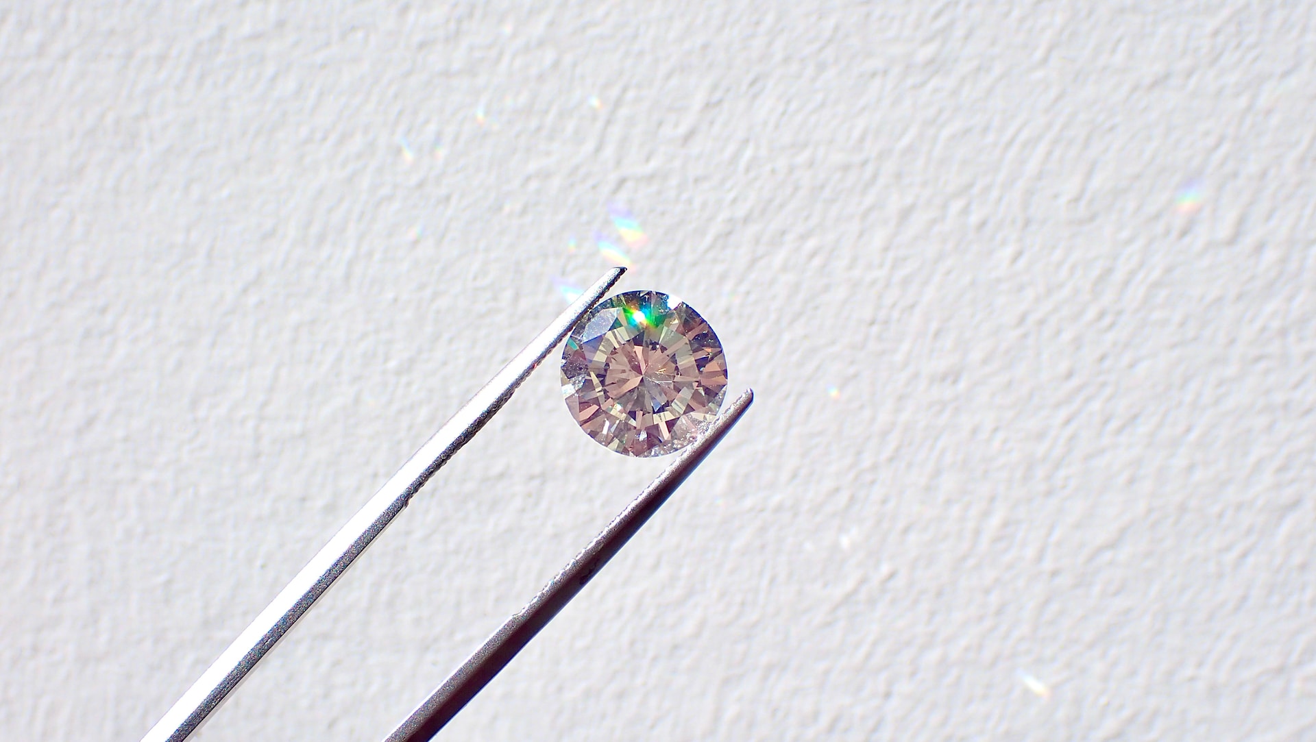 A close-up of a diamond held between jeweler’s tweezers against a white wall.