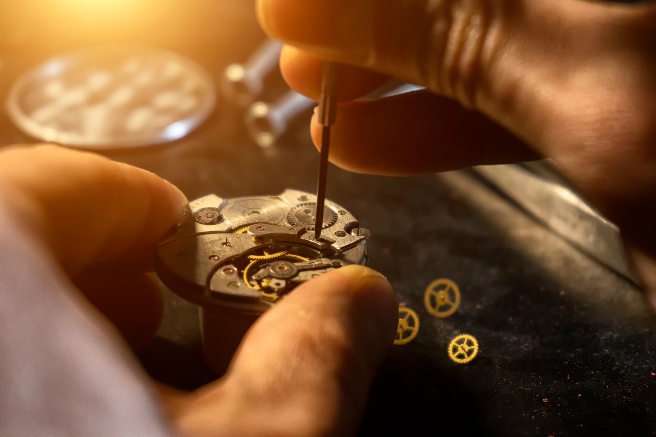 Close up of hands carefully repairing a watch mechanism with a precision tool, surrounded by tiny gears on a work surface