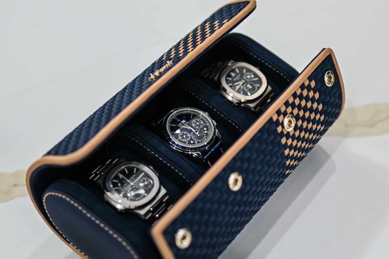 A navy blue and beige checkered watch case displays three high-end wristwatches with metal bands