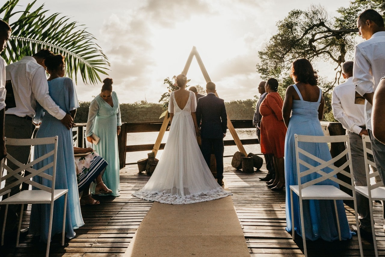 A couple stands facing a triangular arch during an outdoor wedding ceremony with guests in colorful attire watch, and the sun sets in the background.