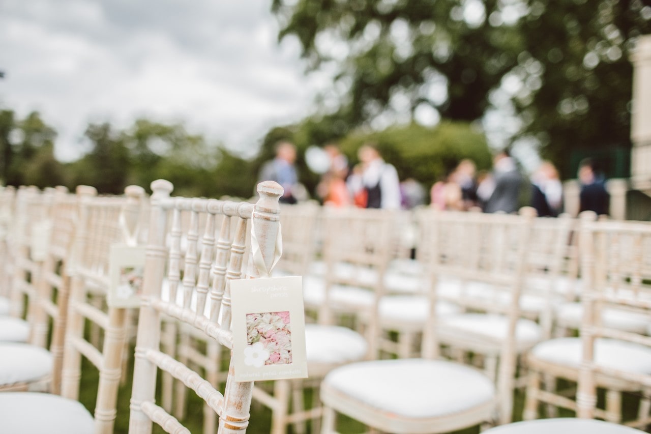 Rows of white chairs with floral cards attached are arranged outdoors for a wedding, while in the blurred background, people gather