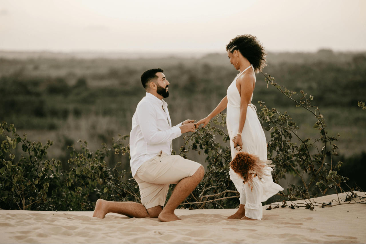 A man kneels on sandy ground, proposing to a woman in a flowing white dress, while she holds flowers against a blurred natural landscape at sunset.